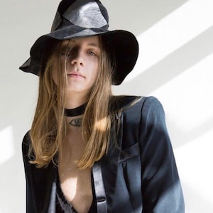 May include: A person with long brown hair wearing a black wide-brimmed hat, a black blazer, and a black choker with an eye design.