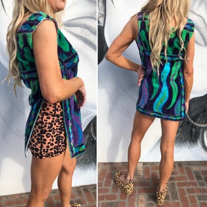 May include: A sleeveless mini dress with a vibrant abstract pattern in shades of green, blue, and purple. The dress has a V-neckline and a fitted silhouette. The model is wearing the dress with platform shoes.