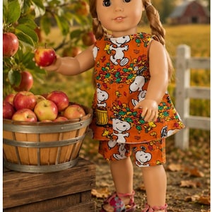 May include: A doll with light brown hair in pigtails wearing an orange sleeveless top and matching shorts featuring a Snoopy and autumn leaf print. The doll wears pink sandals and holds a red apple near a wooden basket filled with red apples.