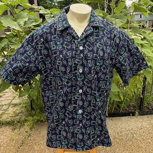 May include: A short-sleeved button-down shirt in dark navy blue featuring an all-over print of colorful retro video game consoles and handheld controllers in neon shades of pink, blue, and green, with a classic collar and white buttons.
