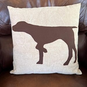 May include: A square decorative pillow with a brown dog silhouette on a cream-colored background. The pillow is placed on a brown leather couch. The dog silhouette is a German Shorthaired Pointer.