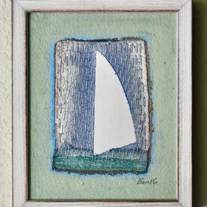 Puede incluir: Obra de arte enmarcada que representa un velero con una vela blanca sobre un fondo azul y gris. La obra está enmarcada en un marco de madera blanco y tiene un borde verde claro. La firma del artista, "Ban G", es visible en la esquina inferior derecha.
