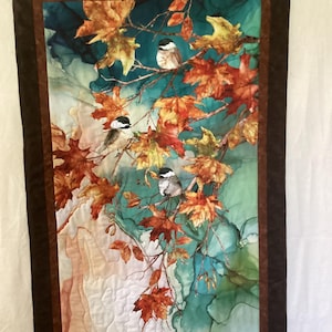 May include: A quilted wall hanging featuring a vibrant autumn scene. The artwork depicts several small birds perched on branches surrounded by colorful fall leaves in shades of orange, yellow, and brown against a teal and white background. The piece is framed with a dark brown border.
