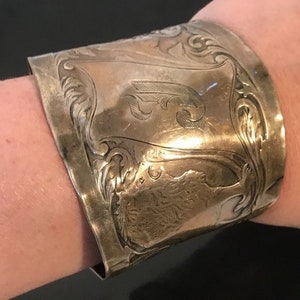 May include: A silver cuff bracelet with an ornate design and a monogrammed initial. The bracelet is wide and has a rounded shape.