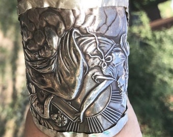 May include: A silver cuff bracelet with a detailed embossed design of a figure holding a book. The design is intricate and appears to be a mythological or historical scene.