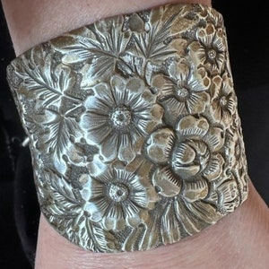 Puede incluir: Un brazalete de plata con un diseño floral. El brazalete está hecho de metal y tiene un patrón floral detallado.