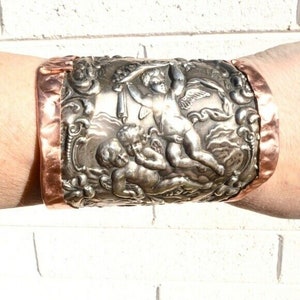 May include: A silver cuff bracelet with a detailed design of cherubs and floral motifs. The bracelet is accented with a copper band.