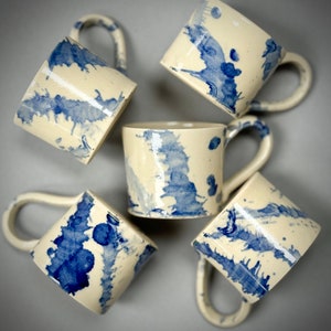 May include: Five white ceramic mugs with blue watercolor-like designs. The mugs have rounded handles and are arranged in a circle on a gray background.