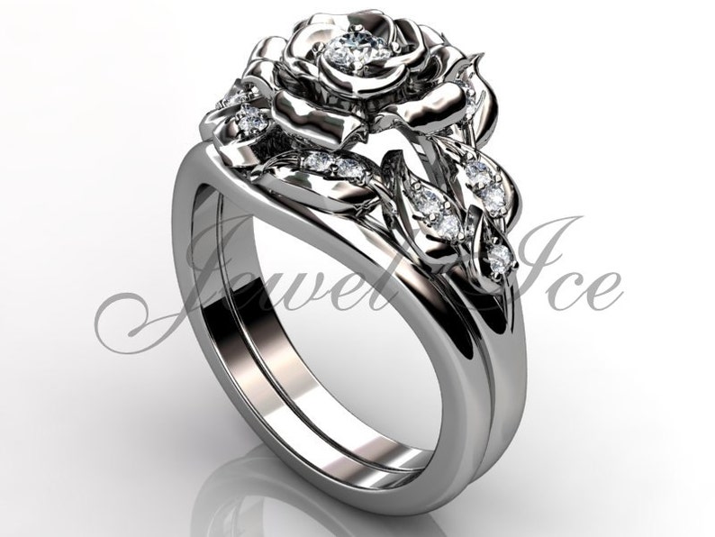 Puede incluir: Un anillo de compromiso de plata en forma de rosa con un diamante central y diamantes m&aacute;s peque&ntilde;os en la banda y las hojas. El anillo est&aacute; sobre un fondo blanco.