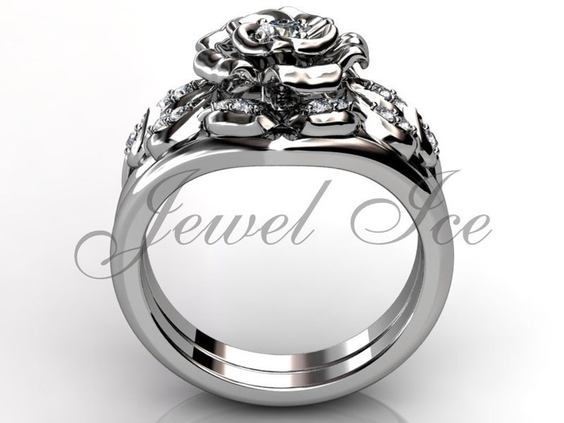 Puede incluir: Un anillo de compromiso de oro blanco en forma de rosa con un diamante central y diamantes m&aacute;s peque&ntilde;os en la banda. El anillo est&aacute; sobre un fondo blanco.
