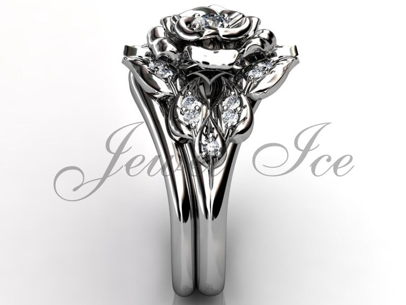 Puede incluir: Un anillo de compromiso de oro blanco en forma de rosa con un solo diamante en el centro de la rosa y diamantes m&aacute;s peque&ntilde;os en las hojas. El anillo est&aacute; sobre un fondo blanco.
