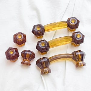 May include: Three amber-colored arched drawer pulls and three matching hexagonal knobs. Each piece features a translucent yellow center with dark brown, faceted ends. The hardware has a smooth, glossy finish and a distinct geometric shape.