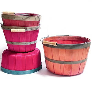 May include: A collection of four round wooden baskets featuring vertical slats and metal rims. Three baskets are stacked vertically in a vibrant fuchsia pink, while one separate basket stands to the right in a gradient of orange and pink hues.