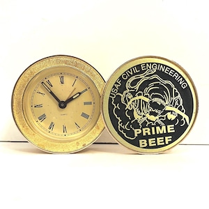 May include: A gold-toned quartz clock with Roman numerals and black hands, next to a round, gold-rimmed emblem. The emblem features a black illustration and the words "USAF CIVIL ENGINEERING" and "PRIME BEEF."