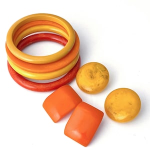 May include: A collection of vintage jewelry pieces in shades of orange and yellow. The set includes several bangles in varying shades of orange and yellow, along with two round and two square-shaped pieces. The pieces are arranged on a white surface.
