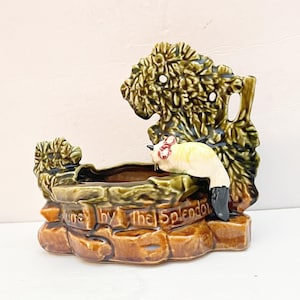 May include: A ceramic planter shaped like a wishing well with a glossy brown stone-textured base and a dark green leafy backdrop. A small cream-colored cat with a red bow sits on the rim. The text "...by the Splendor" is embossed on the front.