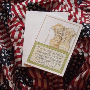 Op de afbeelding: Een wenskaart met een aquarel illustratie van gevechtslaarzen, vergezeld van een witte envelop. De kaart bevat tekst binnen een groene rand, tegen een achtergrond van een Amerikaanse vlag.