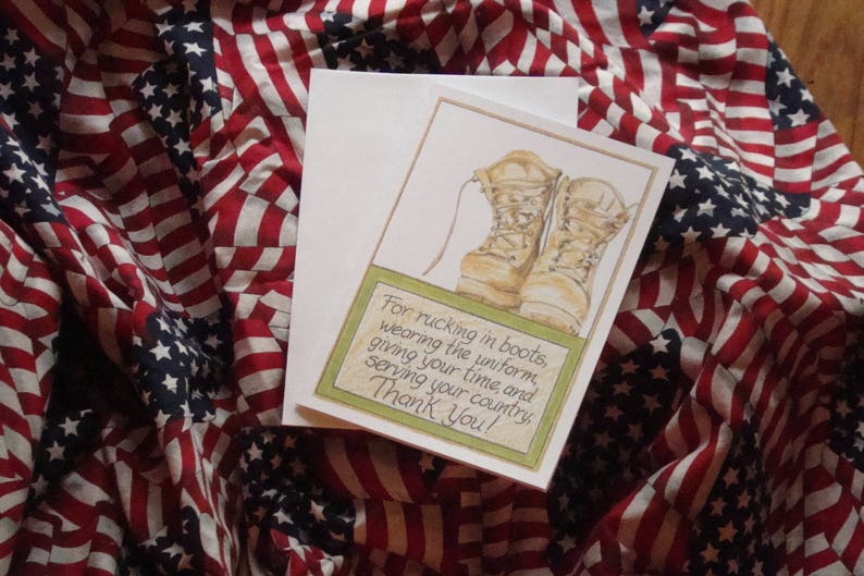 Op de afbeelding: Een wenskaart met een aquarel illustratie van gevechtslaarzen, vergezeld van een witte envelop. De kaart bevat tekst binnen een groene rand, tegen een achtergrond van een Amerikaanse vlag.