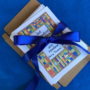 May include: A stack of white cards with colorful book illustrations and the quote "Not all readers are leaders, (Harry Truman)" is tied with a blue satin ribbon. The cards are in a brown cardboard envelope, set against a blue background.