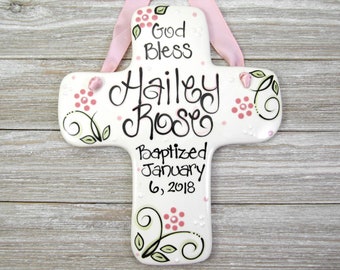 May include: White ceramic cross with pink and green floral designs. The cross is decorated with white dots and pink flowers. The text on the cross reads "God Bless Hailey Rose Baptized January 6, 2018".