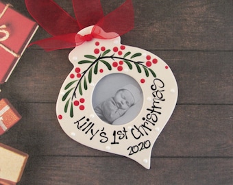 May include: A white, teardrop-shaped ceramic Christmas ornament featuring a central circular photo frame, hand-painted green holly leaves with red berries, and the black handwritten text "Lilly's 1st Christmas 2020" with a red ribbon bow.