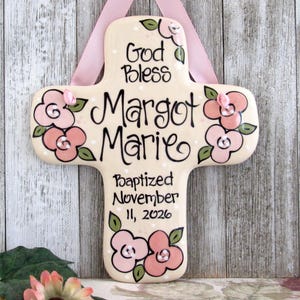 May include: A cream-colored ceramic cross wall hanging featuring hand-painted pink flowers with green leaves and black lettering that reads God Bless Margot Marie Baptized November 11, 2026. A light pink satin ribbon is attached for hanging.