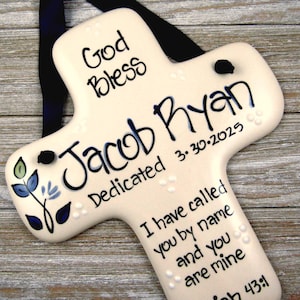 May include: A cream-colored ceramic cross wall hanging featuring a black ribbon for suspension. The surface is hand-painted with the text God Bless, Jacob Ryan, Dedicated 3.30.2025, and I have called you by name and you are mine Isaiah 43:1.