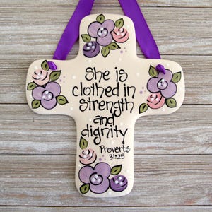 May include: A cream-colored ceramic cross featuring hand-painted purple and pink flowers with green leaves. The text reads, "She is clothed in strength and dignity Proverbs 31:25" in black script. A purple ribbon is attached for hanging.