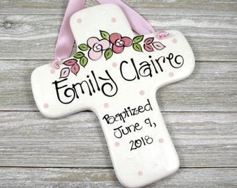 May include: White ceramic cross with pink polka dots and a floral design. The cross is decorated with pink flowers and green leaves. The text "Emily Claire" and "Baptized June 9, 2018" is written on the cross.