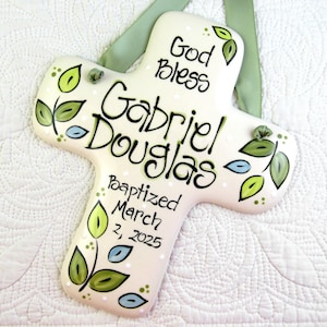 May include: A white ceramic cross with green and blue leaves painted on it. The cross has the text "God Bless Gabriel Douglas Baptized March 2, 2025" written on it.