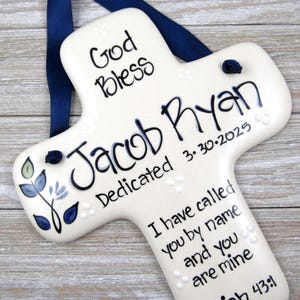May include: A white ceramic cross with blue and black accents. The cross is decorated with a floral design and the text "God Bless Jacob Ryan Dedicated 3-30-2025 I have called you by name and you are mine 43:1".