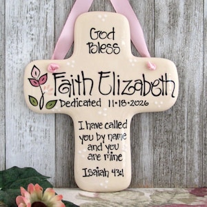 May include: A cream-colored ceramic cross wall hanging with a pink ribbon hanger. It features the text God Bless, Faith Elizabeth, Dedicated 11-18-2026, and the Bible verse I have called you by name and you are mine Isaiah 43:1, with a floral motif.
