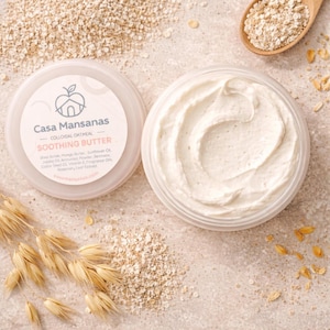 Könnte beinhalten: Geöffnetes Glas Casa Mansanas Soothing Butter mit kolloidalem Hafer. Die weiße Creme ist strukturiert und gewirbelt. Der Deckel liegt neben dem Glas mit Markennamen und Produktbeschreibung. Hafer und ein Holzlöffel mit Hafer befinden sich im Hintergrund.