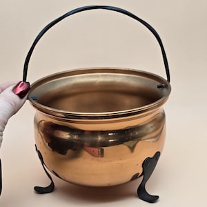 May include: A copper-colored cauldron with a black handle and three black feet. The cauldron has a rounded body and a wide opening. The handle arches over the top. The cauldron is likely used for decorative purposes.