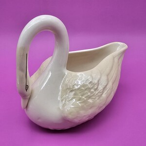 May include: A white ceramic swan creamer with a long, curved neck and a detailed wing design. The swan's head is gracefully arched, and the body features textured feather detailing. The creamer is set against a purple background.