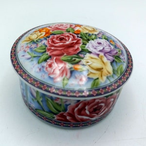 以下が含まれることがあります： 花柄の丸い磁器製の小物入れ。蓋には、薄い青色の背景に、ピンク、黄色、紫のバラが緑の葉と共に集まっている。箱の底は、大きなピンクのバラ1つと、同様の花柄のデザインになっている。