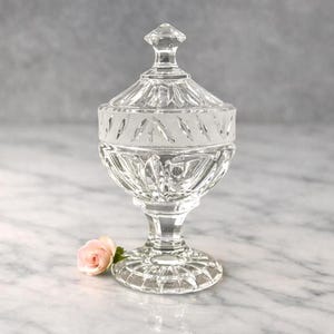 May include: A clear glass candy dish with a lid, featuring a faceted finial, a frosted band with leaf-like patterns, and a pedestal base. The dish has a cut glass design with vertical ridges and a circular foot, accompanied by a small pink rose.