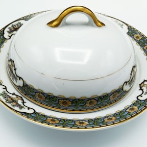 May include: A white porcelain butter dish with a gold-trimmed lid and a floral design. The dish has a blue and green floral pattern with yellow flowers.