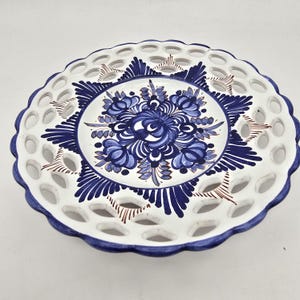 May include: White ceramic plate with a blue floral design in the center and a scalloped edge. The plate has a pierced design around the rim and is trimmed with blue paint. A decorative piece for serving or display.