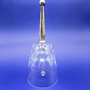 May include: A clear glass bell with a faceted design and a gold-colored handle. A small, clear crystal ball hangs inside the bell. The bell is set against a blue background.