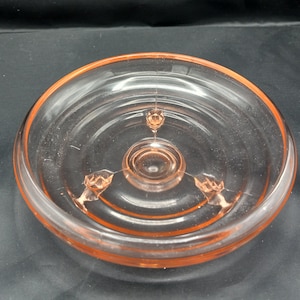 May include: A clear glass pedestal dish with a pink hue and three feet. The dish has a scalloped edge and a raised center.
