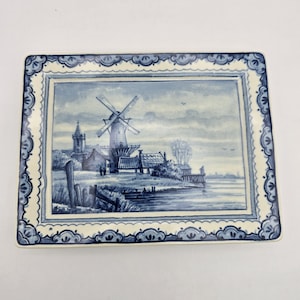 May include: Rectangular ceramic plate featuring a blue and white Dutch windmill scene. The plate has a decorative border with a scalloped edge. The windmill is the focal point of the landscape, with a church and houses in the background.