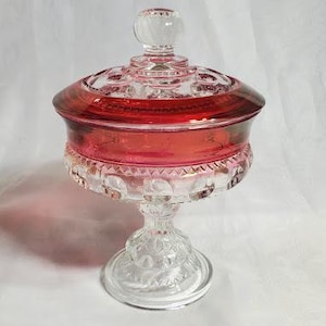May include: A vintage glass candy dish with a clear pedestal base and a red glass lid with a clear knob. The dish has a raised, beaded rim and a clear glass base.