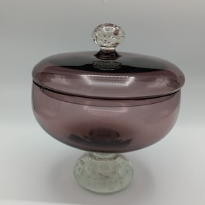 May include: A decorative glass jar with a lid. The jar is a deep purple color and sits on a clear, textured base. The lid is a darker shade of purple with a clear, round knob on top. The jar is designed for storage or display.