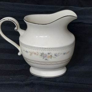 May include: A white ceramic creamer with a silver handle and a floral design. The creamer has a delicate floral pattern in shades of pink, blue, and yellow.