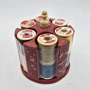 May include: A circular maroon plastic thread spool holder containing six spools of Clark's Boilfast Mercerized thread. Each spool features a white label with red text reading Clark's 35 Yds or size 50. A small wooden knob sits at the center top.