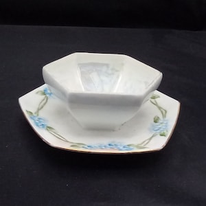 May include: White porcelain sauce boat with a geometric octagonal shape and a matching saucer. The saucer has a delicate floral design with blue flowers and green leaves.