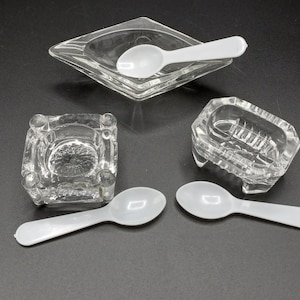 May include: Four clear glass salt cellars with four white plastic spoons. The salt cellars are square, rectangular, and diamond shaped.