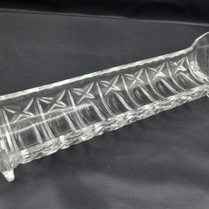 May include: Clear glass serving dish with a long, rectangular shape and starburst pattern. The dish has a curved end and a textured rim. The clear glass allows for visibility of the contents.