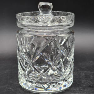 Puede incluir: Un tarro de cristal transparente con tapa. El tarro tiene un patrón de corte de diamante y una tapa redonda con un pequeño pomo. El tarro cilíndrico está vacío. El fondo es negro.